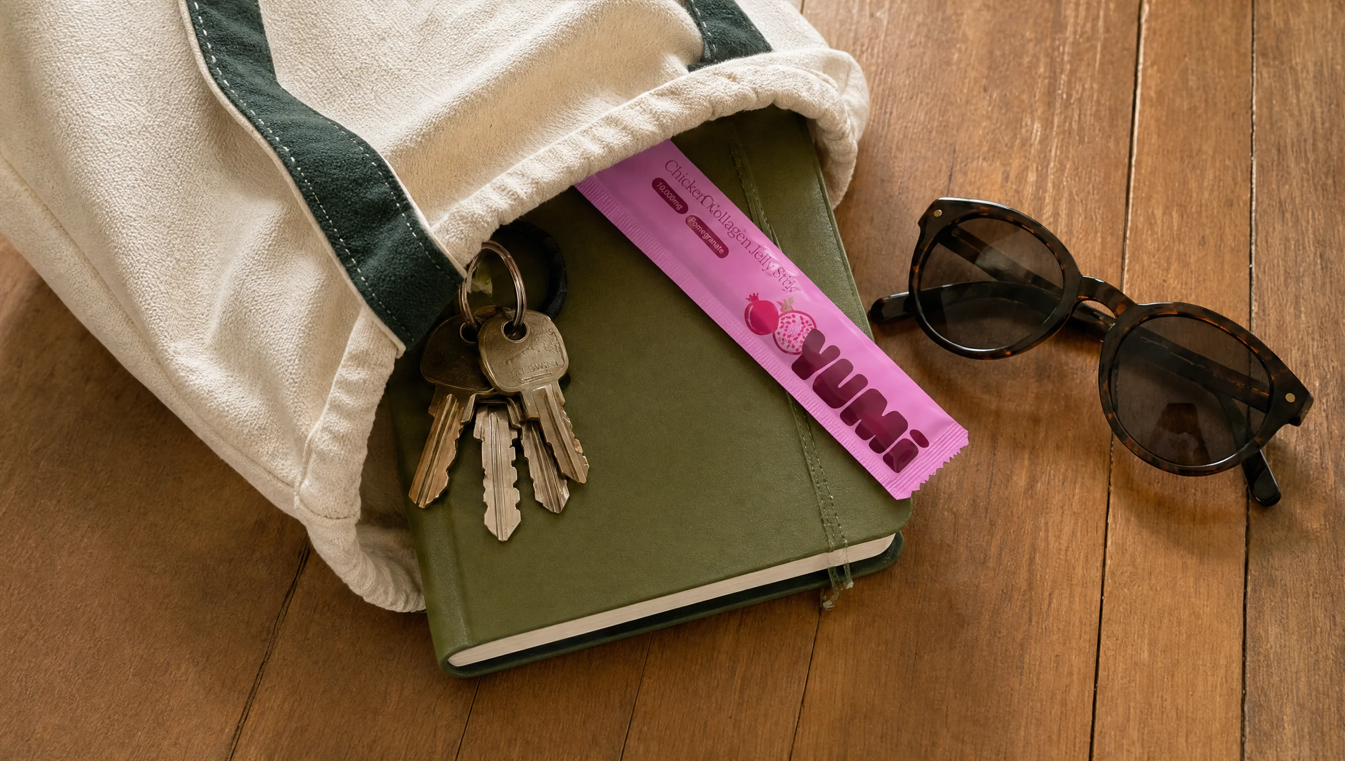 A pink Yumi jelly stick tucked into a tote bag with a journal, keys, and sunglasses on a wood floor.